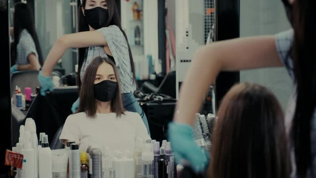 Woman client in mask sits down into chair to have haircut and stylist puts on cape at mirror with replicated reflection in salon