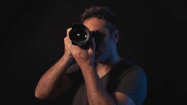 Portrait of a young professoinal male photographer panning the camera working at the studio. Isolated artist with black background. Freelance working with a mirrorless DSLR camera