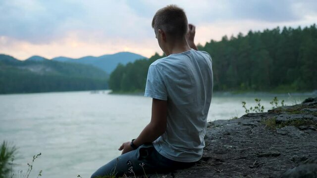 The teenager sits on the banks of the river on a cliff. He throws rocks into the water