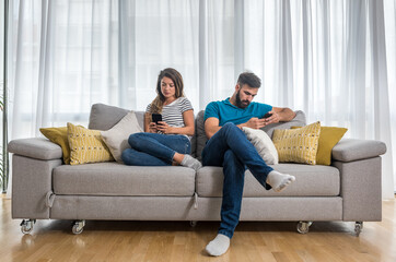 Obraz premium Young couple sitting on the sofa in the living room and ignoring each other by surfing and text messaging on their smartphones, relationship difficulties and issues concept