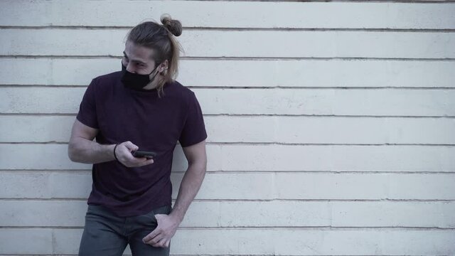 Long Haired Caucasian Man Wearing Black Mask And Shirt Using Smartphone While Standing On The Streetside By The Wall - medium shot