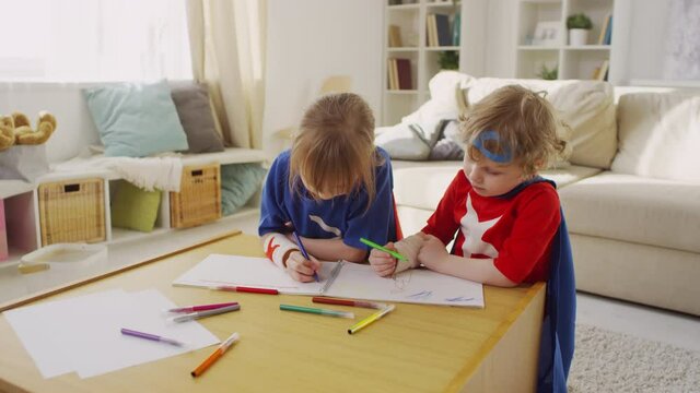 Medium shot of small happy children in superhero costumes drawing with colourful markers at table in cozy living room