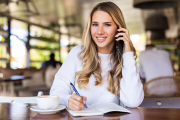 Cheerful young woman talk phone conversation on smartphone doing order and making notes of information about delivery. Positive hipster girl calling on mobile phone sitting with coffee