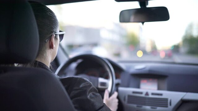 Person driving car in city. Traffic passing by. Female taxi cab or rideshare driver. Commute or road trip. Rental vehicle. Inside back view with steering wheel and cockpit. Transportation concept.