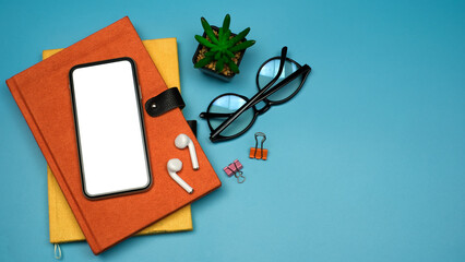 Flat lay, Workspace back to school and education concept on blue table desk with blank screen phone on stack of book, earphone and eyeglass, green plant, Top view with copy space