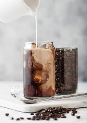 Pouring milk into glass with iced black coffee with glass container of beans and spoon on marbel board and light background.