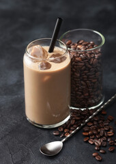 Glass of iced cold coffee and milk with free raw coffee beans in glass container with long spoon on black.