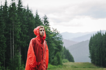 Thoughtful young handsome man in a red waterproof coat is pensively looking aside standing on the mountain hill in the mountain forest. Wet and rainy weather.