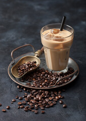 Glass of iced coffee drink with straw on tray with coffee beans and scoop on black background.