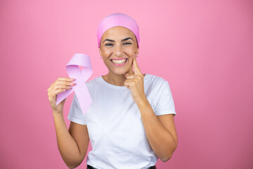 Young beautiful woman wearing pink headscarf holding brest cancer ribbon over isolated pink...