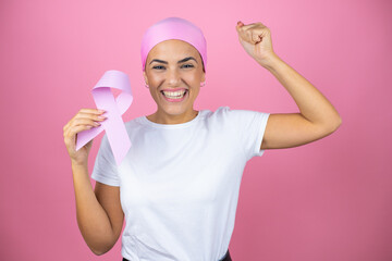 Young beautiful woman wearing pink headscarf holding brest cancer ribbon over isolated pink...