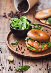 Sandwich with bagel and salmon, cream cheese and wild rocket in bowl and coffee cup with beans on wooden background.