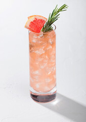 Highball glass of refreshing summer red grapefruit cocktail with ice cubes, fruit slice and rosemary on white background.