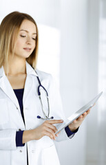 Young woman-doctor is holding a tablet computer in her hands, while standing in a clinic. Portrait of friendly female physician with a stethoscope at work. Perfect medical service in a hospital