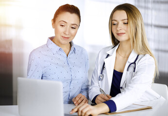 Fototapeta na wymiar Young beautiful woman-doctor and her patient are discussing patient's current health examination, while sitting together at the desk in the sunny cabinet in a clinic. Female physician is writing some