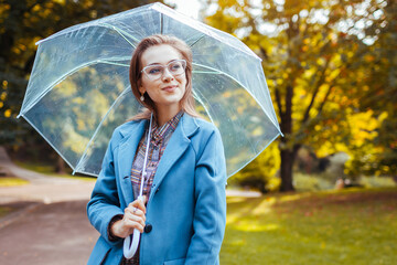 Stylish young woman walking in park with transparent umbrella wearing trendy outfit. Autumn fashion