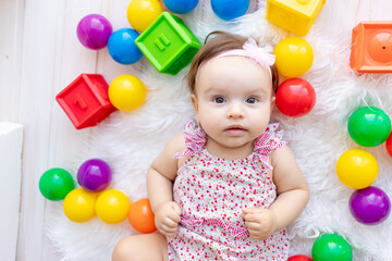a beautiful little baby girl is lying in red clothes on a white Mat among toys balls and cubes