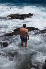 An unrecognizable sturdy man in blue trunks stands in the sea at daytime, facing the challenging strong currents and surf that hit the rocks of the coast.