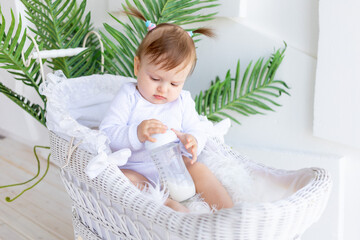 cute little baby girl sitting in a beautiful stroller in a white bodysuit at home and holding a...
