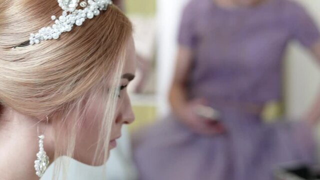 A beautiful young bride with a stylish hairstyle, a diadem on her head and earrings looks at herself in the mirror. Video