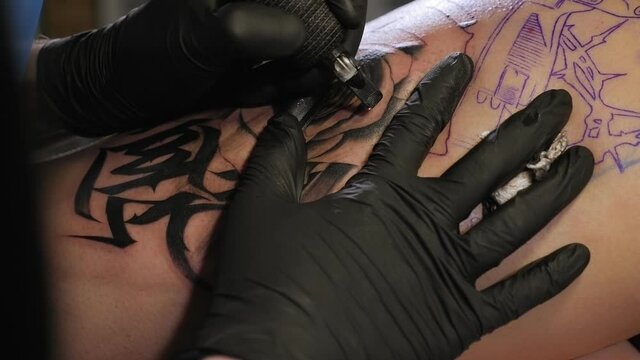Close-up view of professional tattoo artist which making tattoo on the leg of a young woman.