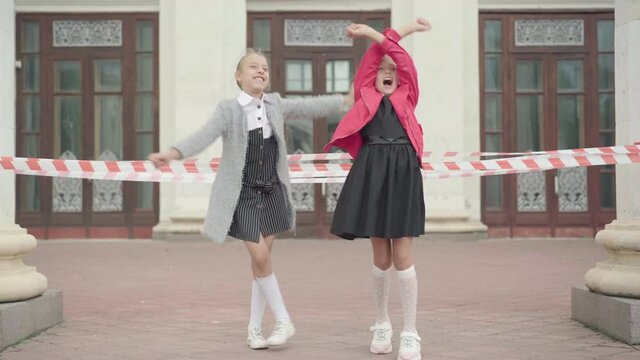 Two joyful carefree schoolgirls rejoicing school closure for Covid-19 pandemic lockdown. Happy Caucasian girls waving to building and jumping with joy. Coronavirus quarantine.