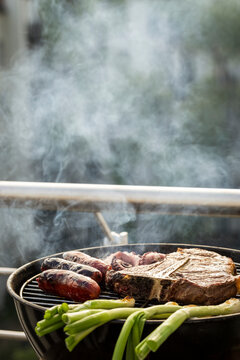 Animal products cooking over hot charcoal grill with rising smoke: sausages, steak, and spring onions during home outdoor barbecue