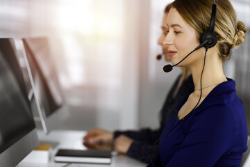 Two busineswomen have conversations with the clients by headsets, while sitting at the desk in a...