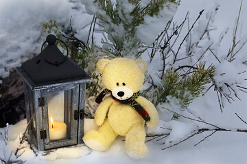 Toy bear with a scarf around his neck sitting in a snowdrift in a snow-covered forest. A lantern with a burning candle stands next to the bear.