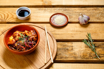 Soby with chicken and sesame seeds in a clay plate on a wooden stand and sticks next to sesame seeds, herbs and soy sauce on a wooden table.