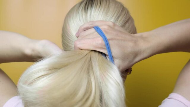 A blond woman styling her hair and making horse tail, ponytail using elastic band against yellow background
