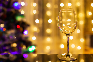 an empty wine glass stands on a black glass table against the background of the Christmas tree and curtains of garlands, the Christmas tree is out of focus