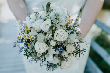 Romantical beautiful bouquet of white roses, chamomile, small blue flowers and bright eared speedwell in brides hands . Woman is wearing beautiful wedding dress. Simple floristic concept.