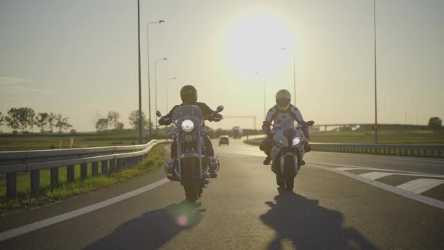Two motorcyclists riding different type of motorcycles fist bumping hands while riding side by side on an empty highway during sunset