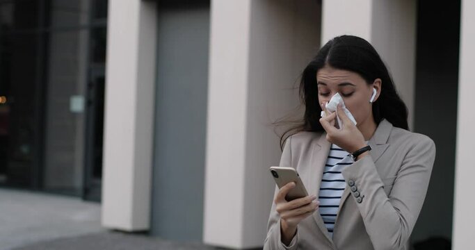 Portrait of young unwell Caucasian woman walking outdoors and using tissue while tapping on smartphone. Sick beautiful female professional with cold and runny nose texting on cellphone. Health concept