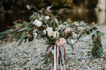 Beautiful big boho bride wedding bouqet of pastel pink roses, lilac eared speedwell, daisy flower and colored tape in small vase is standig on the river background.