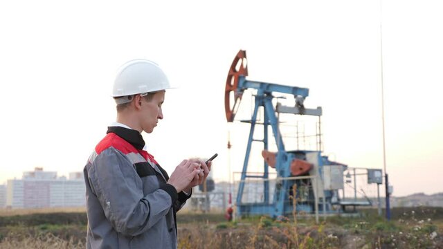 man in a white helmet with a phone against the background of a rocking at an oil well.