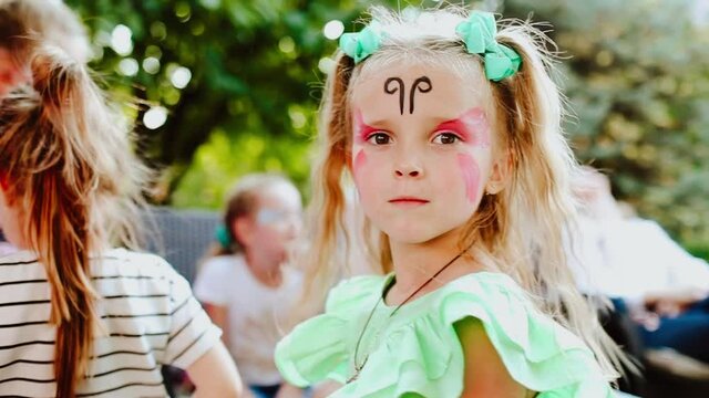 Portrait of young pretty blond girl among the kids with face painting like a butterfly at her face turning around to the camera. Slow motion video.