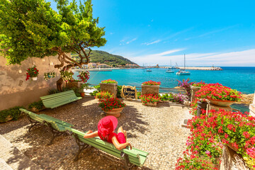 Lifestyle woman in red sitting on bench in Marciana Marina, flowery old district Borgo al Cotone:...
