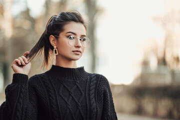 Portrait of young indian woman standing on the street touching her long dark hair, dressed in black...