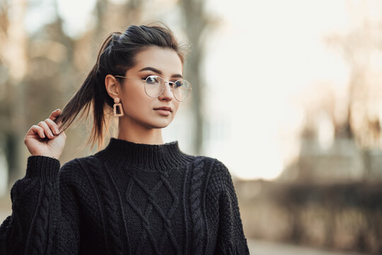 Portrait of young indian woman standing on the street touching her long dark hair, dressed in black pullover with glasses, looking at the camera, image with copy space.