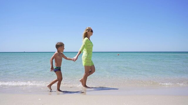 Little boy walks along the sandy shore of the azure sea holding his mother's hand. slow-motion video