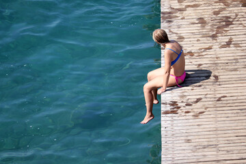Fototapeta premium Girl in bikini sunbathes sitting on wooden pier, top view with free copy space. Beach vacation, relax and leisure by the sea