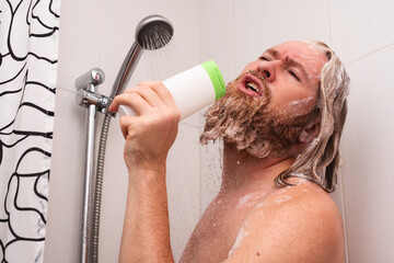 Handsome bearded man singing while taking shower at home using shampoo bottle instead of...