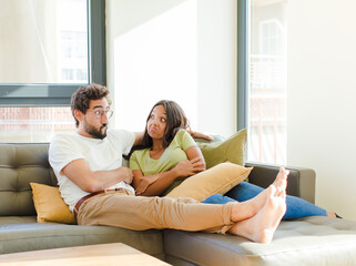 young couple shrugging, feeling confused and uncertain, doubting with arms crossed and puzzled...