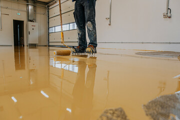 Worker, coating floor with self-leveling epoxy resin in industrial workshop.