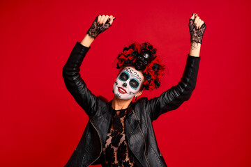 Gir with sugar skull makeup with a wreath of flowers on her head and skull, wearth lace gloves and dances during halloween party isolated on red background.Concept of Halloween or La Calavera Catrina
