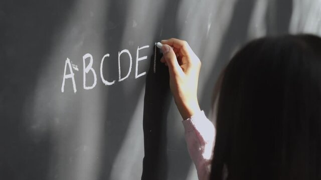 Elementary primary school girl pupil holding chalk writing on blackboard. Child preteen schoolgirl learning english alphabet letters handwriting standing in classroom. Close up back view.