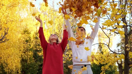 Positive young man and woman in stylish casual clothes throw armfuls of dry bright yellow leaves in sunny autumn park on nice day slow motion