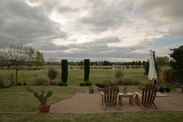 Lifestyle. View of the beautiful green garden with wooden exterior furniture, garden chairs and table, at sunset. 